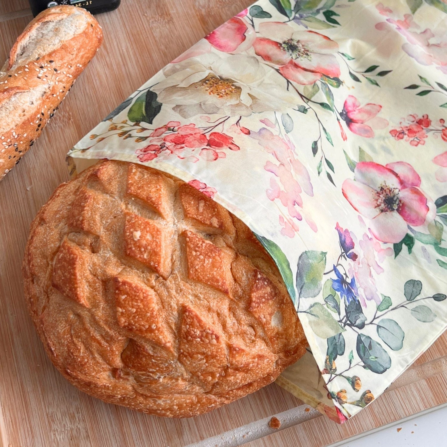 A round artisanal bread loaf wrapped in a floral reusable beeswax bread bag with pink flowers and green leaves, placed on a wooden cutting board with a baguette in the background.