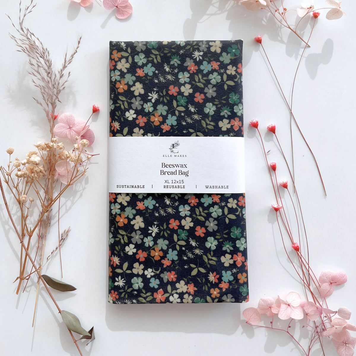 A reusable beeswax bread bag with a dark navy floral print, wrapped in a white paper band . The bag is styled in a flat-lay arrangement with pink flowers and dried botanicals on a white background.