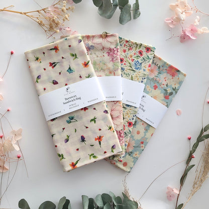 Three folded beeswax bags with floral prints are arranged in a fan shape on a white background with decorative dried flowers and greenery. 