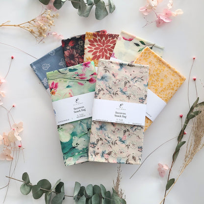Six beeswax snack bags in assorted floral and botanical patterns are arranged in a fan shape on a white background with decorative dried flowers and greenery. 